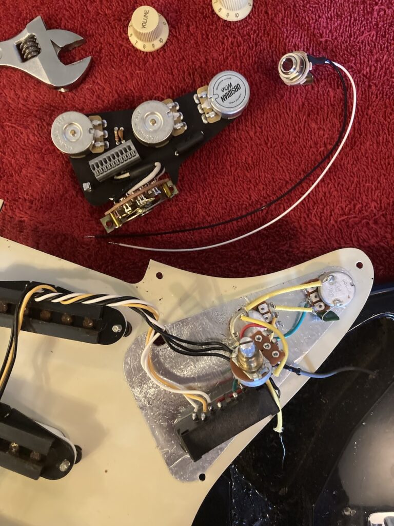 The old potentiometers and switch, still mounted to the underside of the pick-guard, and the new, plate-mounted parts above them, seen from underneath, with the new output jack alongside. Close-up.